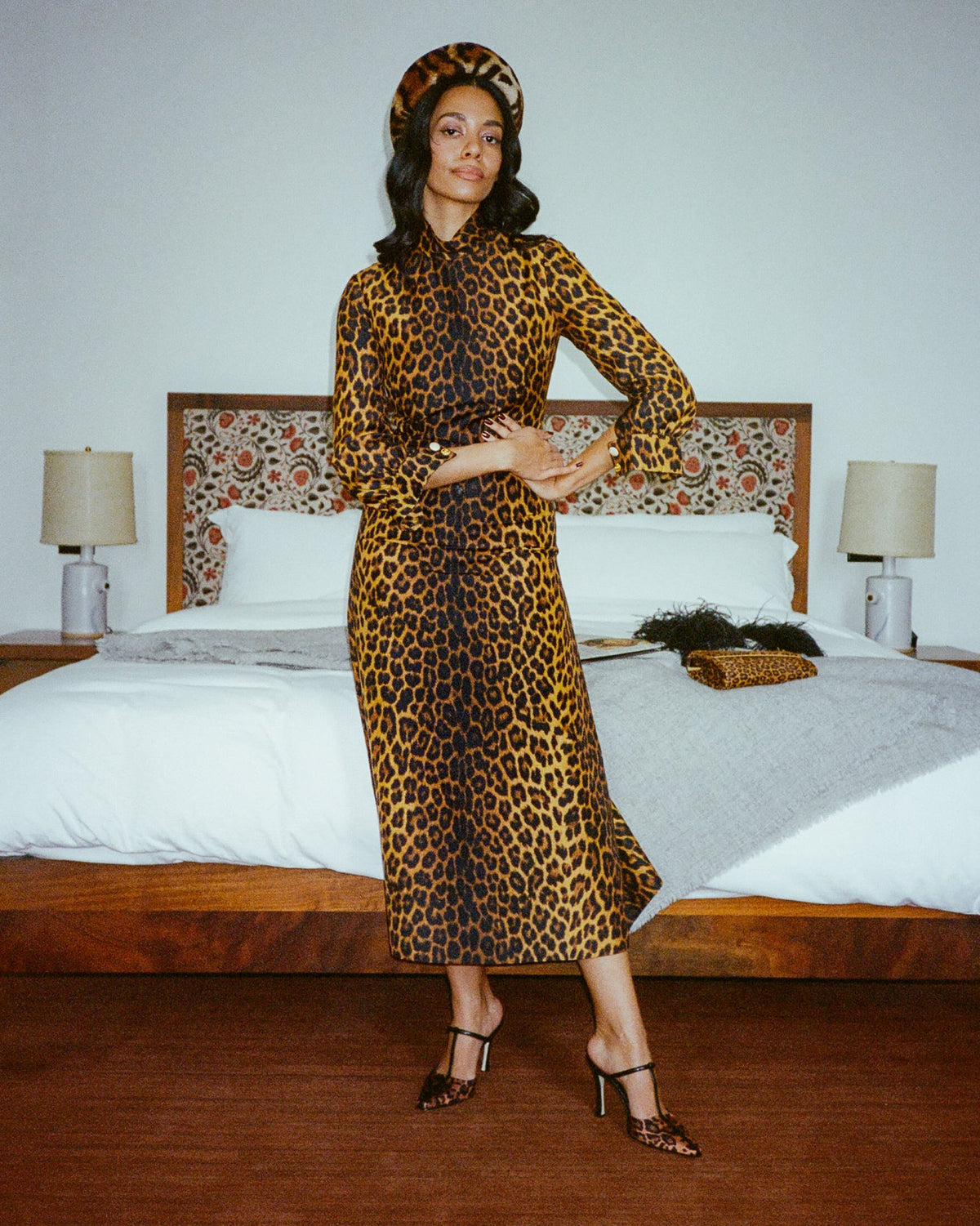 A woman stands in a bedroom wearing a leopard-print midi dress with a matching beret and black strappy heels. The dress has a brown and black animal print on a tan background, and the room features a wooden bed frame, white bedding, and bedside lamps.