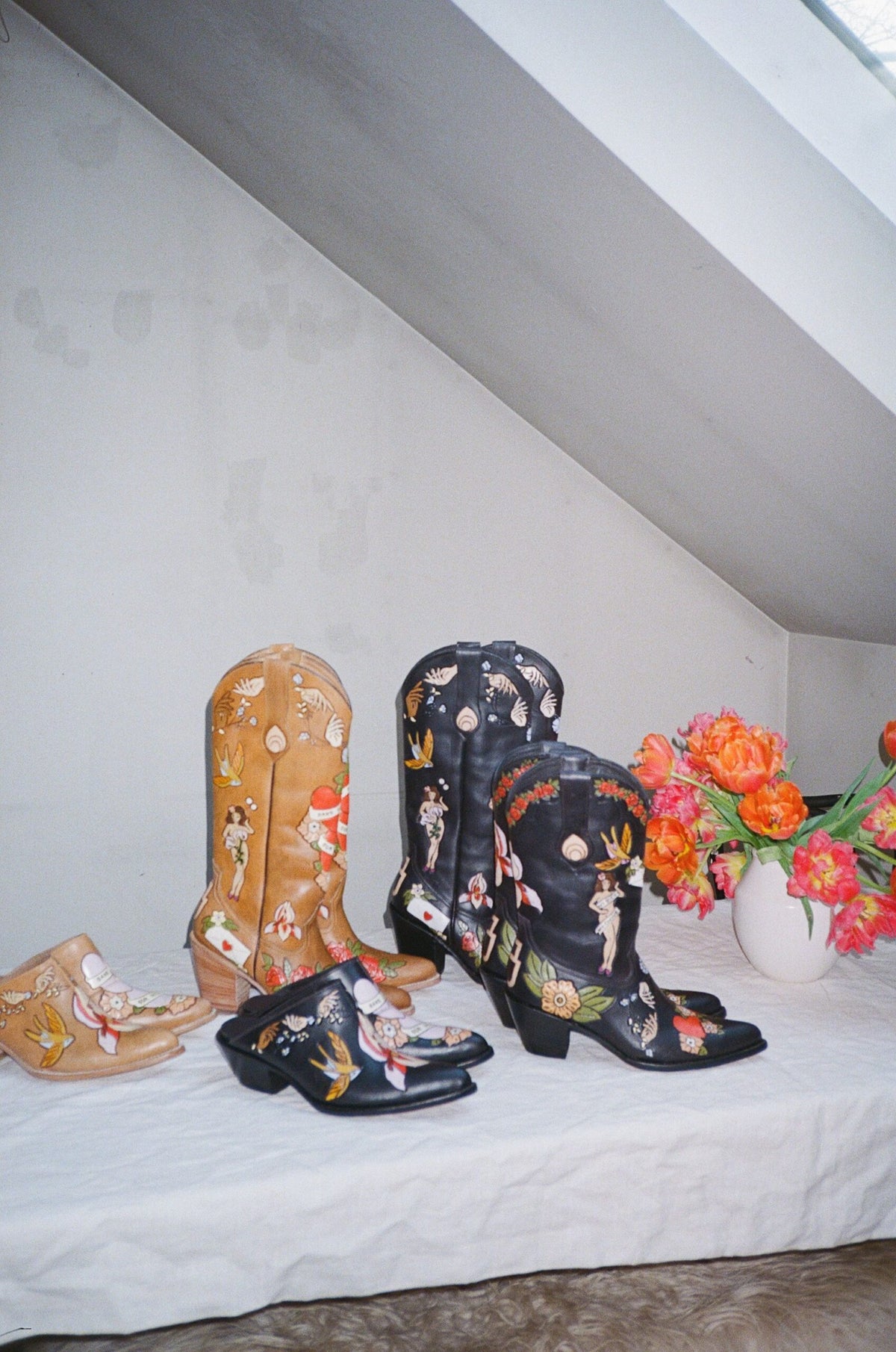 A collection of hand-painted cowboy boots and matching shoes displayed on a white fabric-covered surface in a room with a slanted ceiling. The boots feature colorful floral and western motifs, including one tan boot, one black boot with a multicolor design, and matching black shoes.