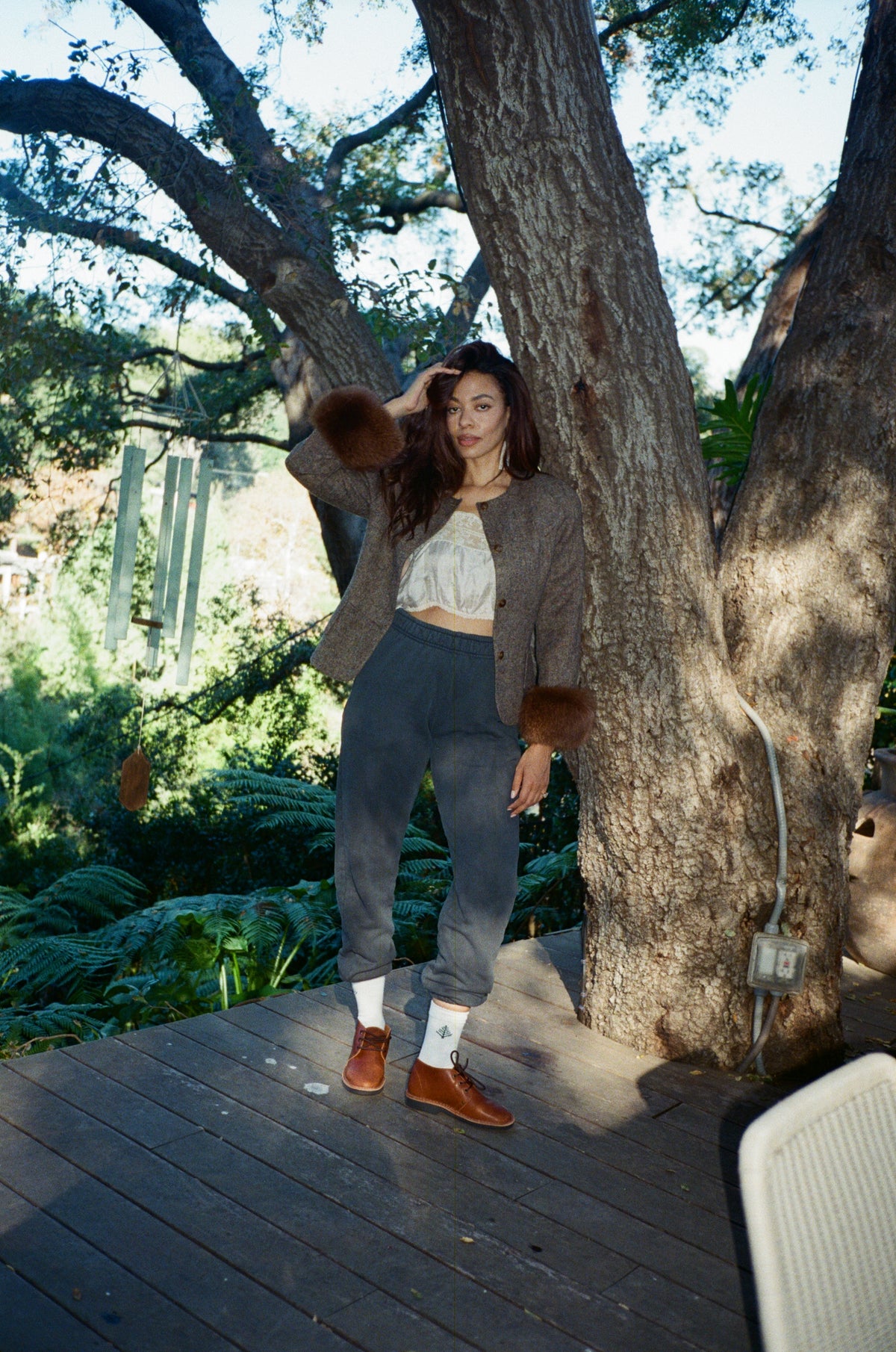 A woman stands on a wooden deck outdoors under a large tree, wearing a brown cardigan, gray pants, white socks, and brown shoes.