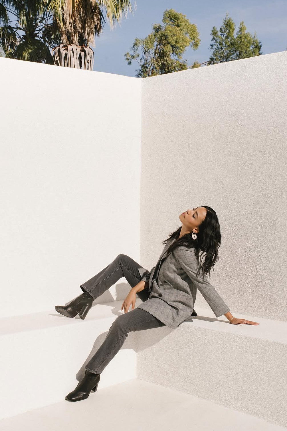 A woman in a gray blazer and matching gray trousers sits on a white architectural corner, looking upward. She is wearing black ankle boots and large white earrings, with white walls, trees, and blue sky visible in the background.