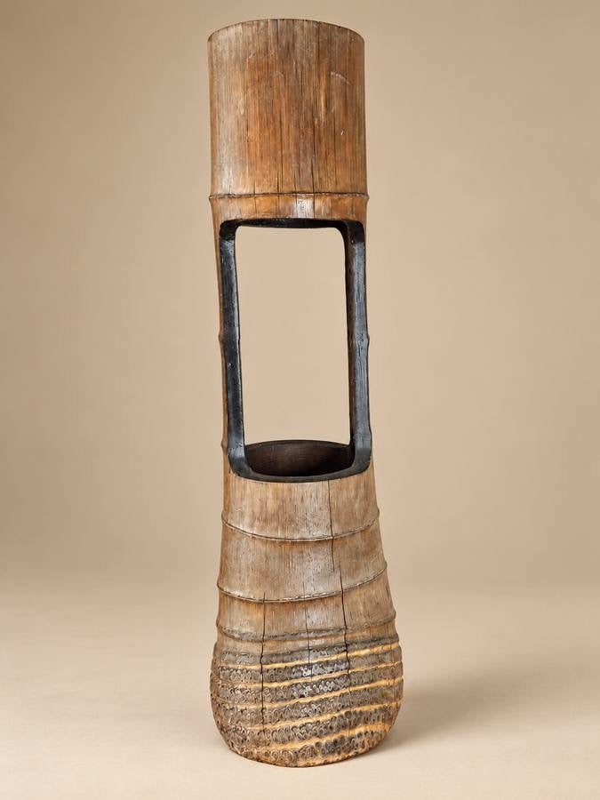 A tall, sculptural wooden decorative piece with a textured natural finish, featuring a cylindrical top, a hollow rectangular middle section, and a flared base, photographed against a solid beige background.