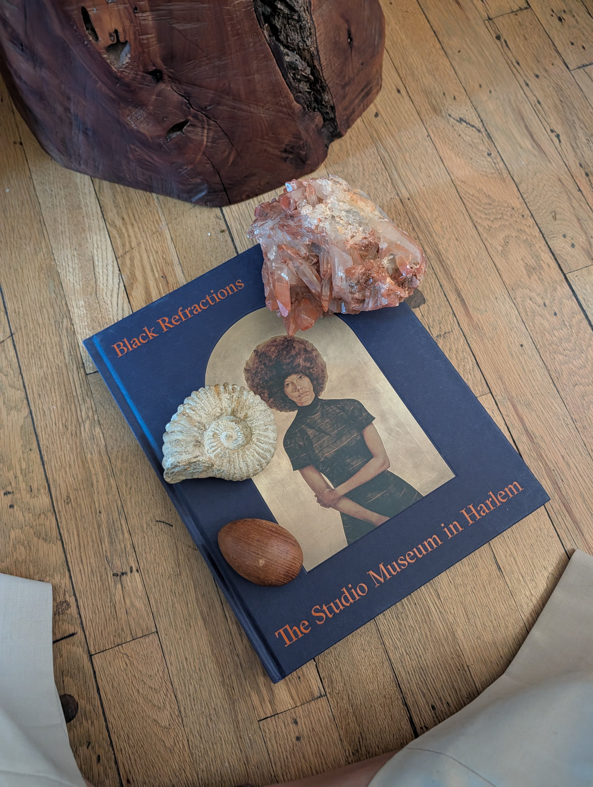 A book titled 'Black Refractions: The Studio Museum in Harlem' lies on a wooden floor, styled with a crystal cluster, a seashell, and a small brown stone. A large dark wood object is partially visible at the top of the frame.