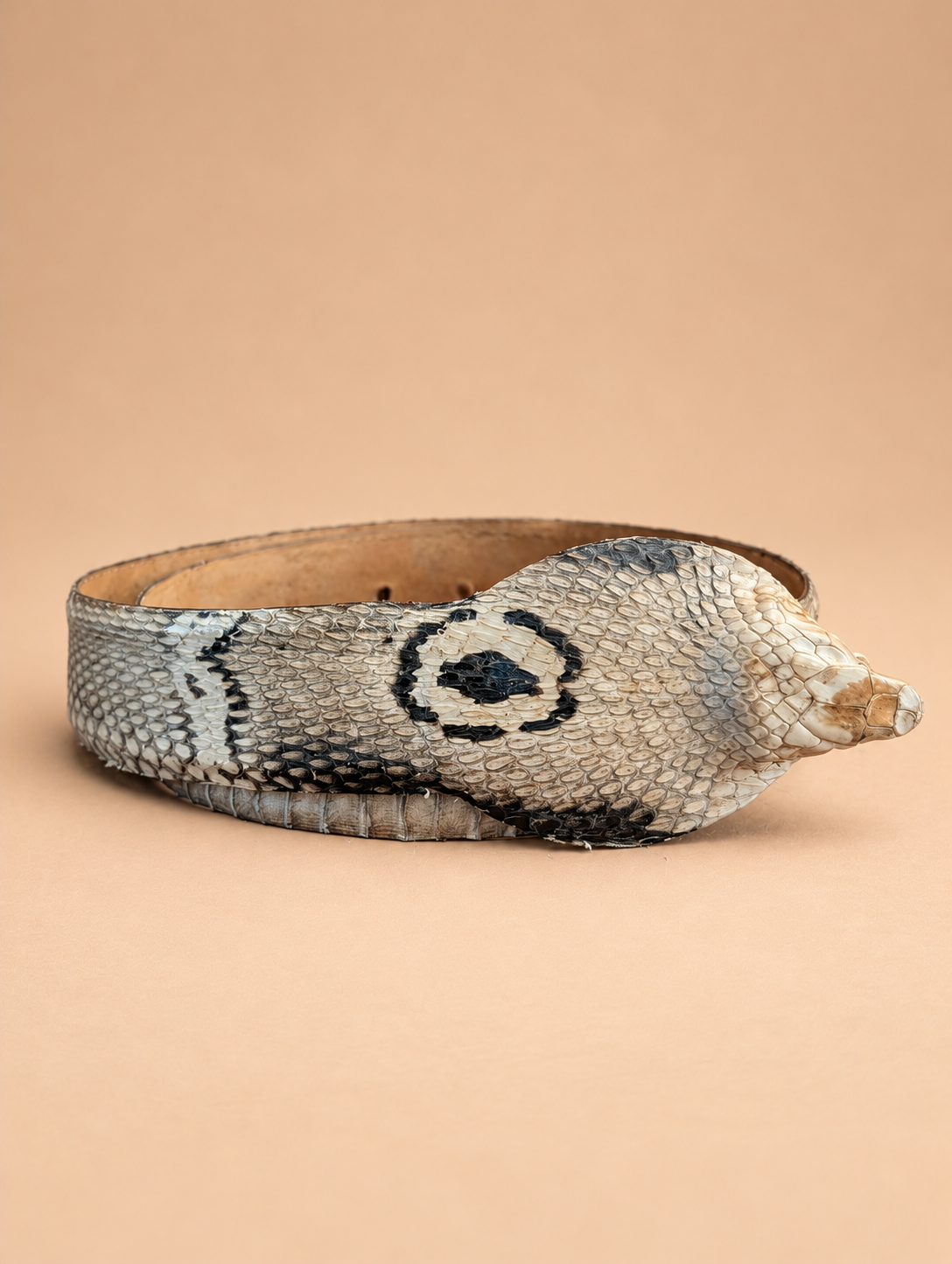 A snake-print leather belt with a textured scale pattern in black, gray, and beige, featuring a circular black eye motif on the front, set against a solid beige background.