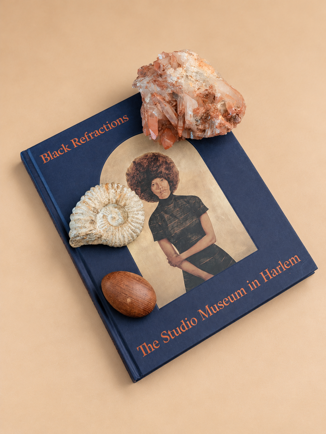 A hardcover book titled 'Black Refractions: The Studio Museum in Harlem' lies on a warm beige background, styled with natural objects including a crystal cluster, a seashell, and a small wooden bead on top.