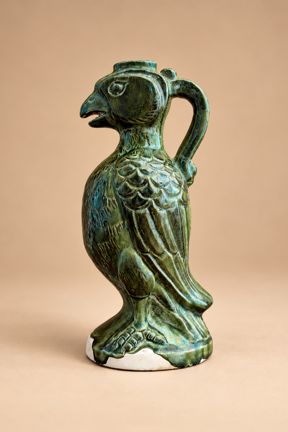 A ceramic bird-shaped pitcher with a green glazed finish, featuring a detailed beak, textured body, curved handle, and small white base, set against a solid beige background.