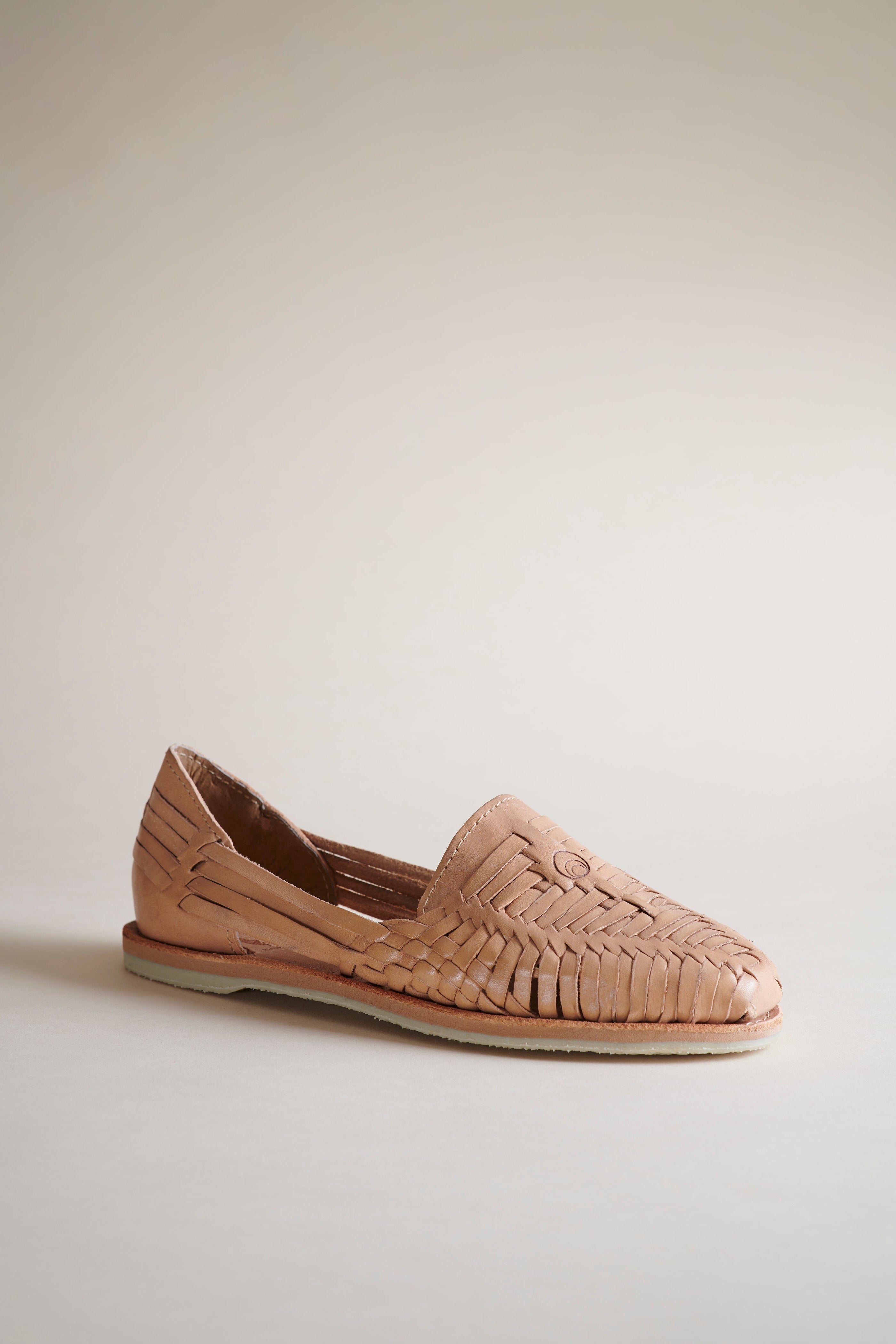 A single woven leather slip-on shoe in a light tan or beige color, shown from a side profile against a neutral, warm off-white background.