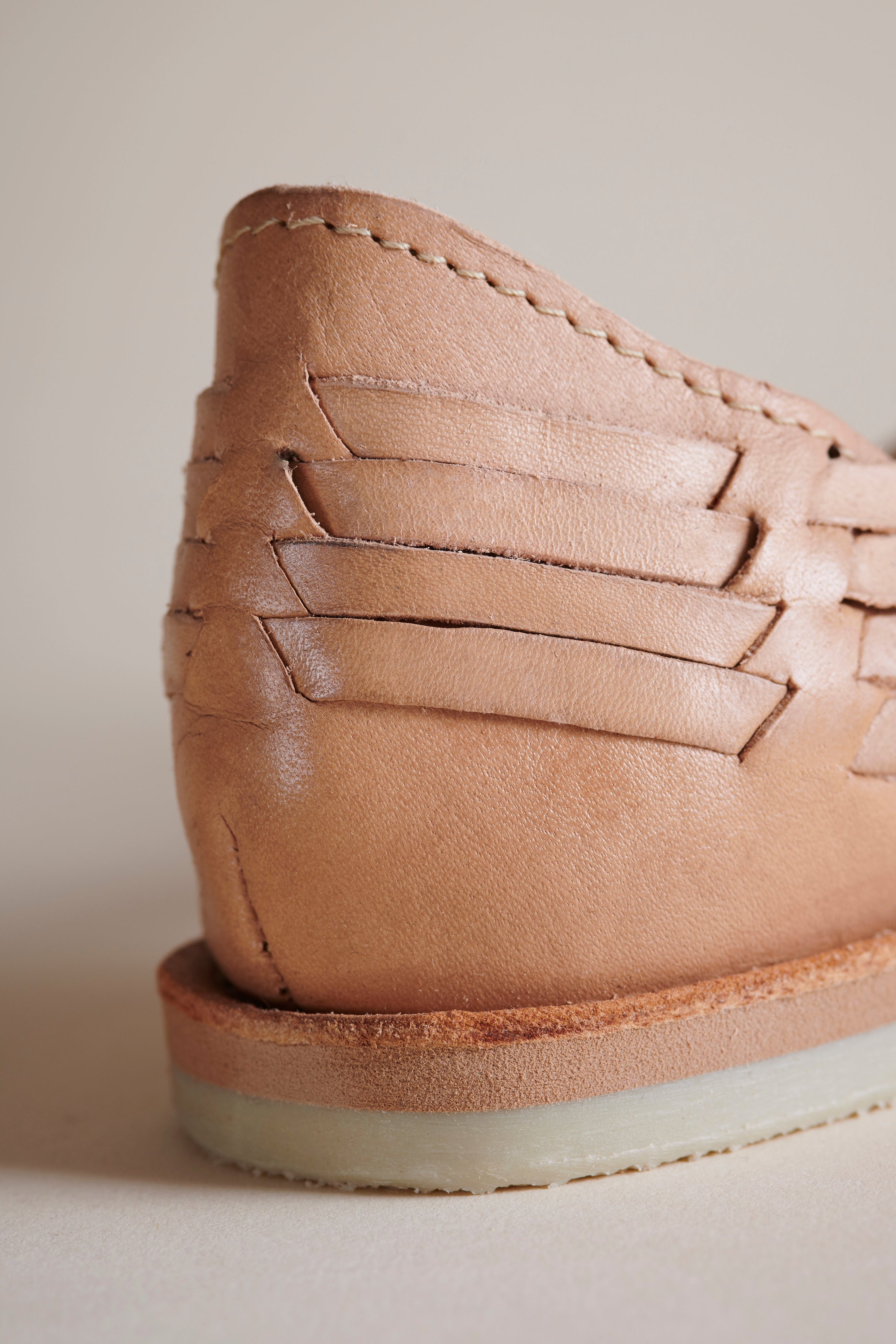 Close-up of the rear quarter of a tan leather shoe with a woven strap detail and a light-colored sole, photographed against a neutral beige background.