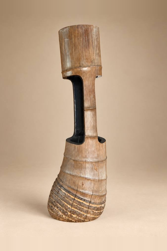 A tall, sculptural wooden vase or decorative vessel with a flared textured base, a narrow neck, and a cylindrical top section, shown against a plain beige studio background.