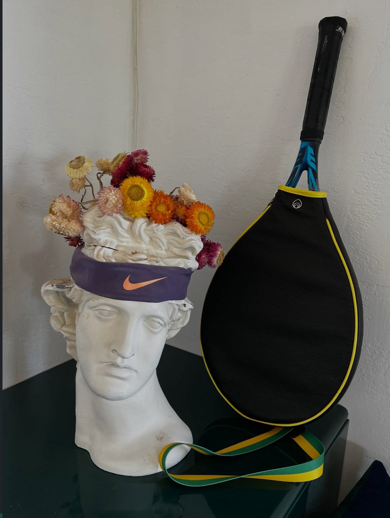 a tennis racquet encased in a leather brother vellies racquet bag, shown displayed on a table leaning against the wall, next to a decorative roman bust statue wearing a flower crown and nike sweatband